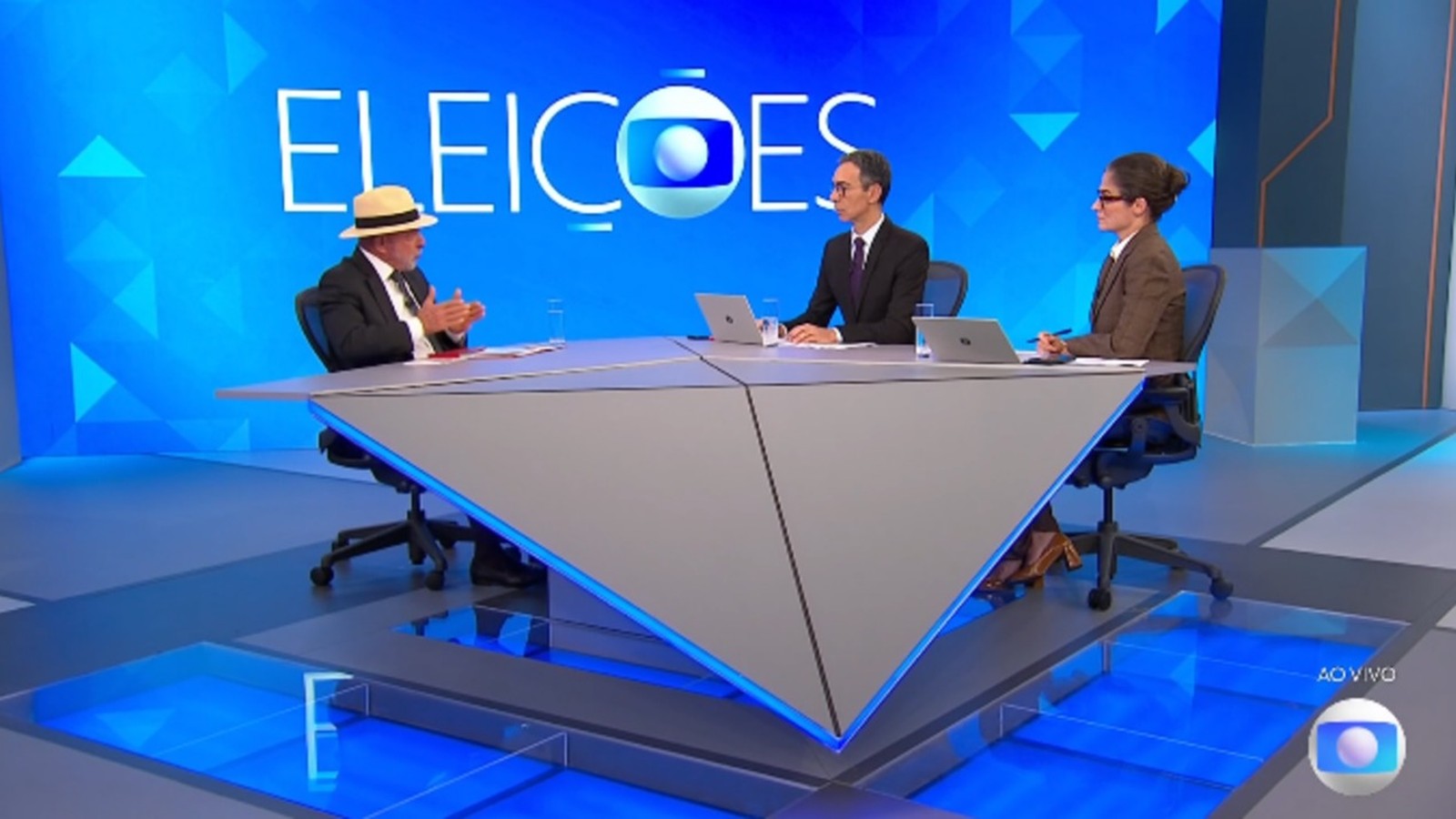 Lula, com chapéu, e dois jornalistas em estúdio de TV, com 'ELEICOES' ao fundo