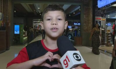 Menino sorridente em shopping faz coração com as mãos para microfone G1