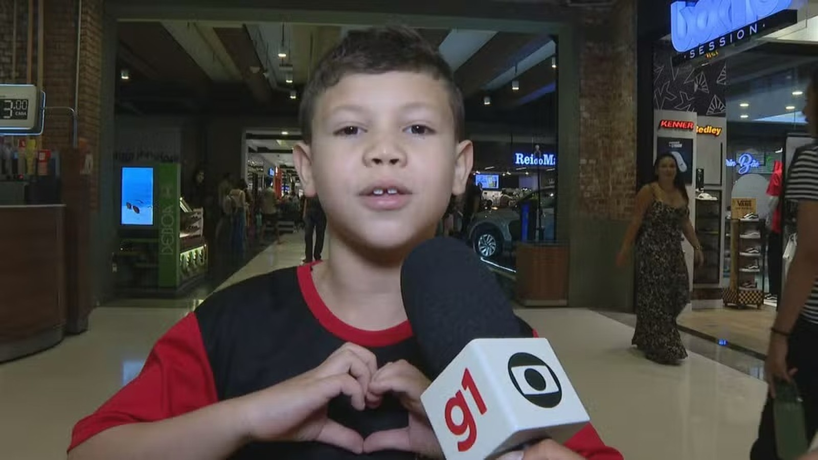 Menino sorridente em shopping faz coração com as mãos para microfone G1