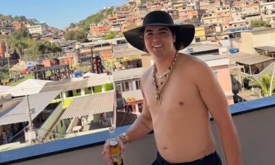 Cantor Nattanzinho Lima, de chapéu, correntes, segurando cerveja, com favela ao fundo