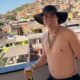 Cantor Nattanzinho Lima, de chapéu, correntes, segurando cerveja, com favela ao fundo