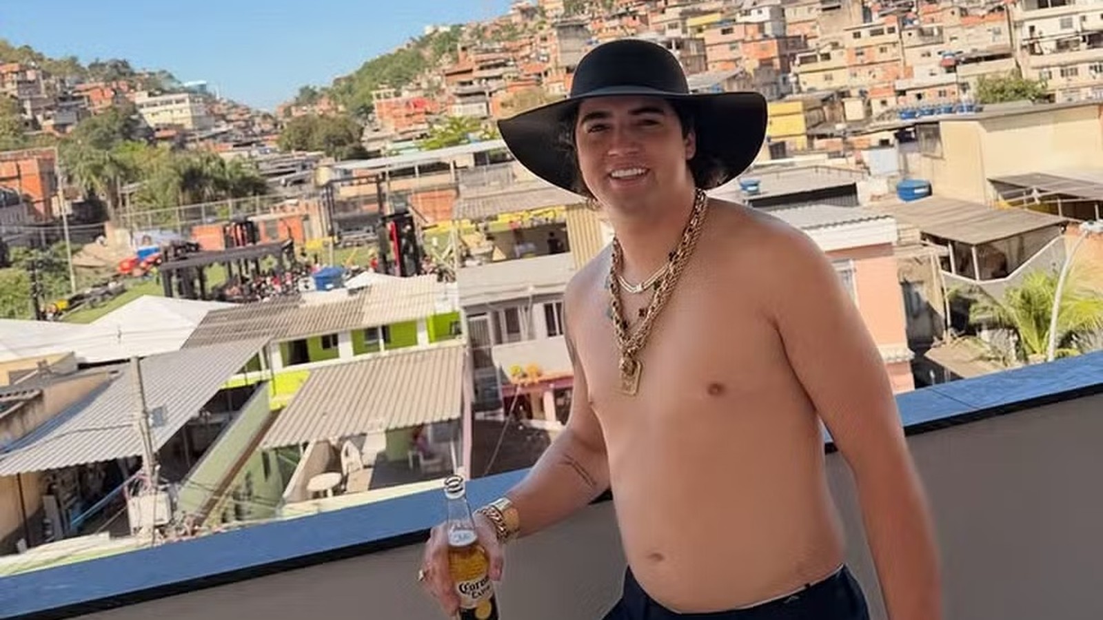 Cantor Nattanzinho Lima, de chapéu, correntes, segurando cerveja, com favela ao fundo