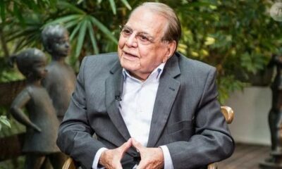 Homem idoso de óculos e terno cinza, sentado, em ambiente com estátuas e folhagem