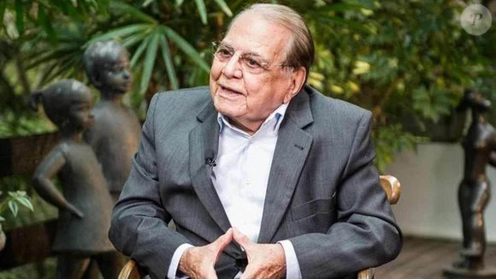 Homem idoso de óculos e terno cinza, sentado, em ambiente com estátuas e folhagem