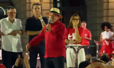 Lula, de camisa vermelha e chapéu, fala ao microfone em palco com outras pessoas