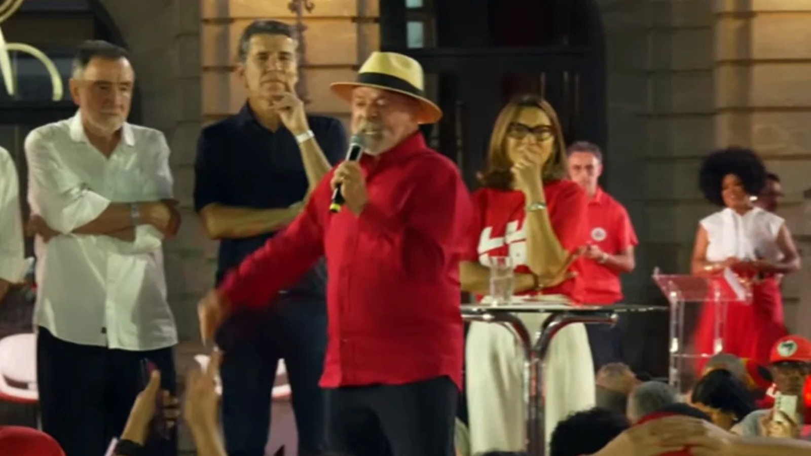 Lula, de camisa vermelha e chapéu, fala ao microfone em palco com outras pessoas