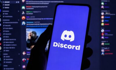 Mão segura smartphone com logo Discord na tela, fundo com interface da plataforma borrada
