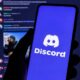 Mão segura smartphone com logo Discord na tela, fundo com interface da plataforma borrada
