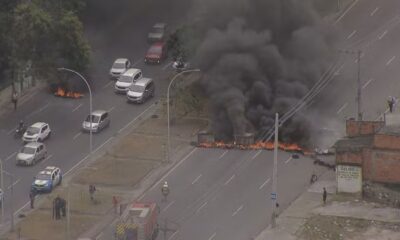 Rua bloqueada por fogo intenso e fumaça preta, carros, polícia e bombeiros no local