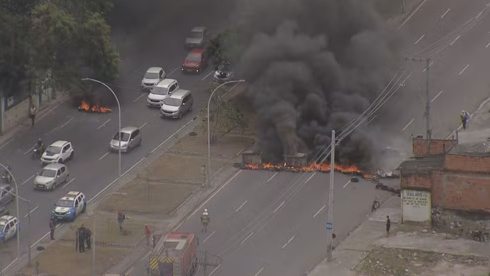 Rua bloqueada por fogo intenso e fumaça preta, carros, polícia e bombeiros no local