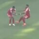Dois jogadores em uniformes vermelhos no campo de futebol, um apoiando o outro