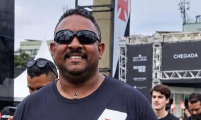 Homem sorrindo, óculos escuros, camisa preta, bigode, em evento ao ar livre com símbolo do Vasco.
