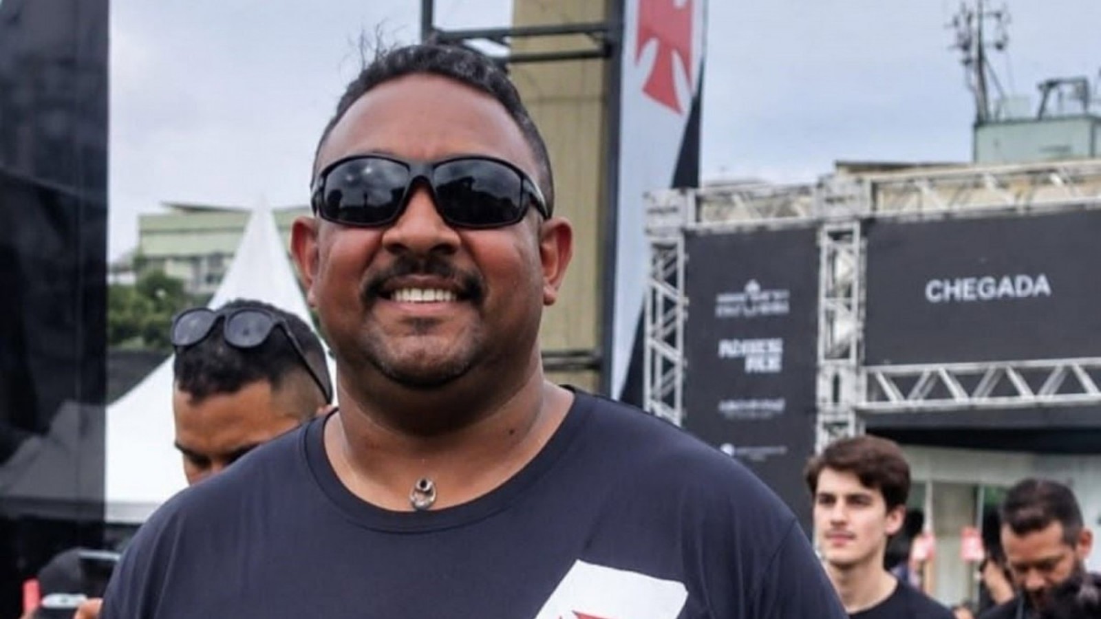 Homem sorrindo, óculos escuros, camisa preta, bigode, em evento ao ar livre com símbolo do Vasco.