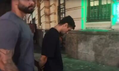 Dois homens caminham em rua, um de barba e outro com camiseta preta e mãos algemadas