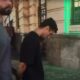 Dois homens caminham em rua, um de barba e outro com camiseta preta e mãos algemadas