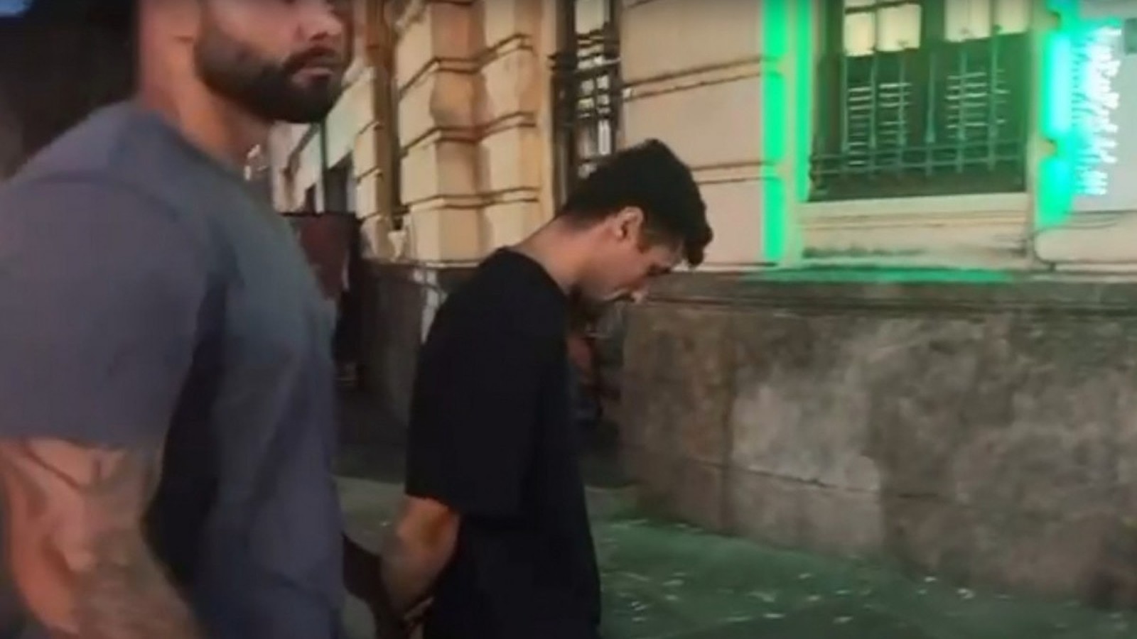 Dois homens caminham em rua, um de barba e outro com camiseta preta e mãos algemadas