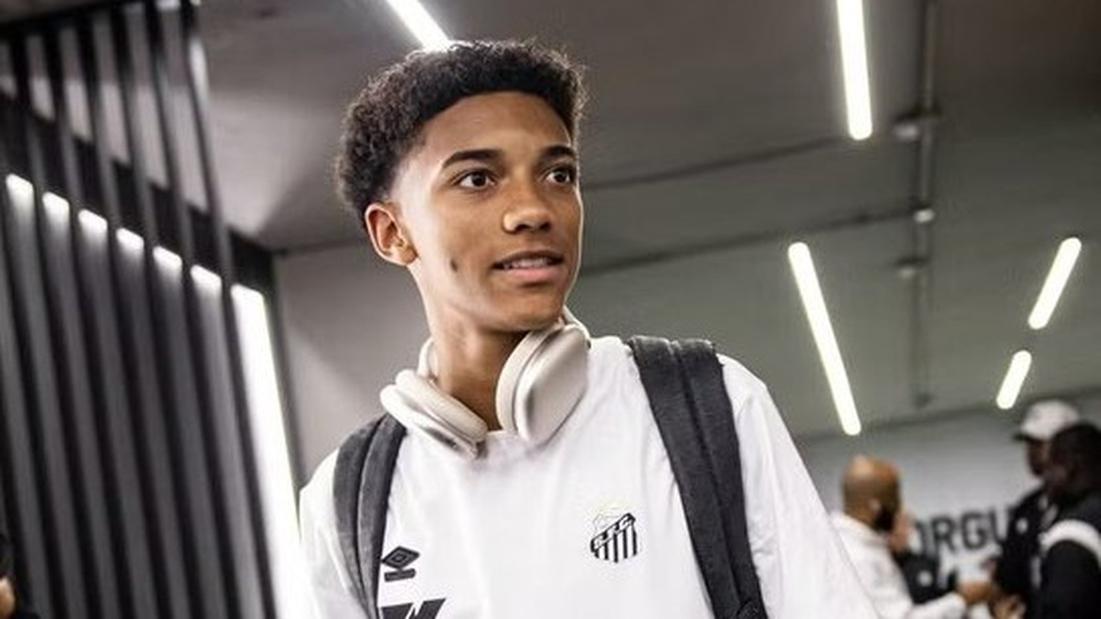 Jovem com cabelo cacheado, camisa do Santos, fones de ouvido no pescoço e mochila