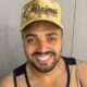 Homem sorridente com barba, bigode e boné dourado escrito "Circo Maximus" olhando para frente