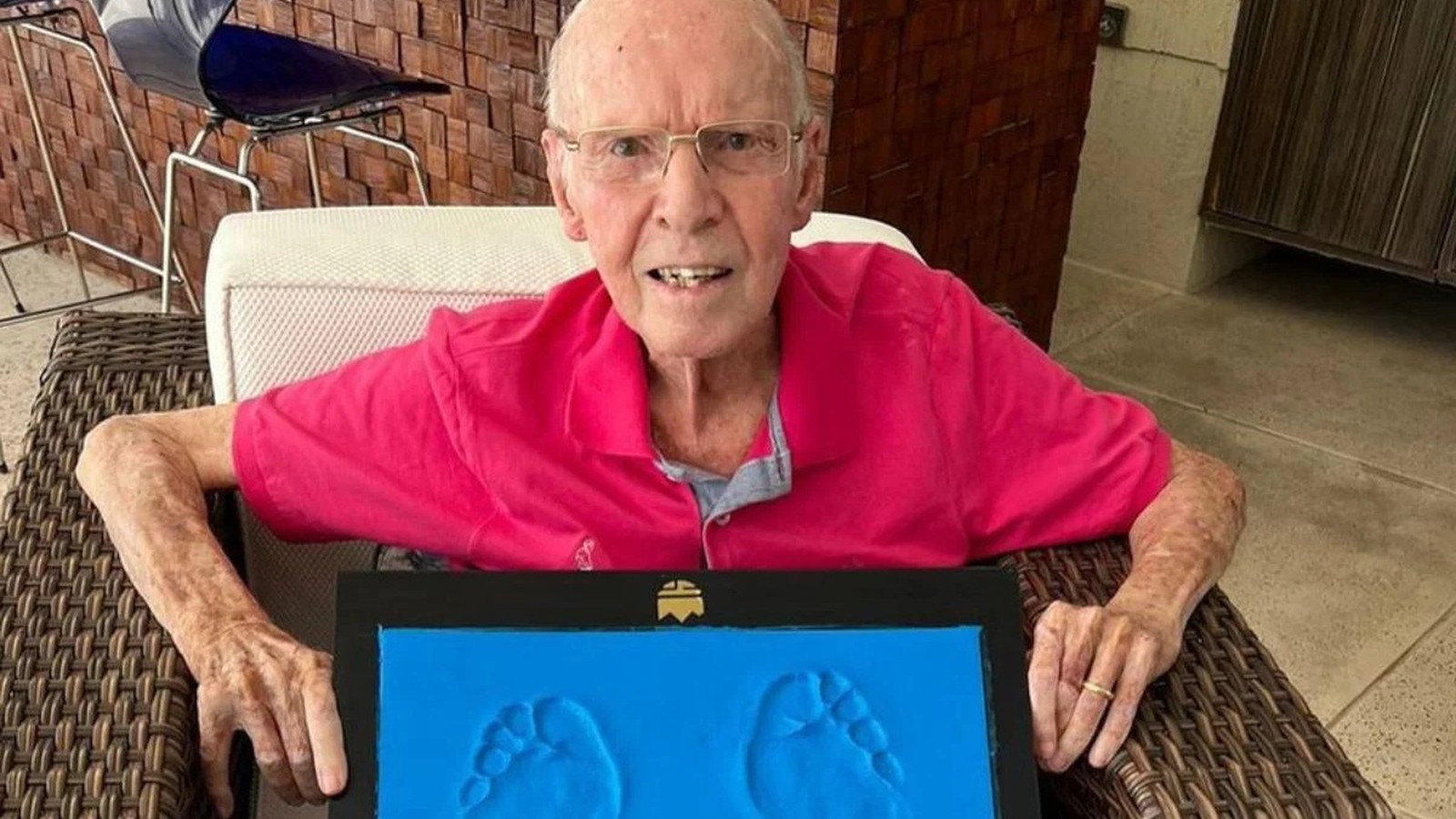 Homem idoso de óculos e camisa rosa segura quadro com pegadas azuis de bebê