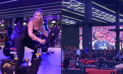 Mulher em bicicleta ergométrica e projeção de Vini Jr. em tela grande na academia