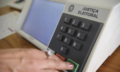 Mão pressiona botão "CONFIRMA" em urna eletrônica da Justiça Eleitoral, com teclado numérico