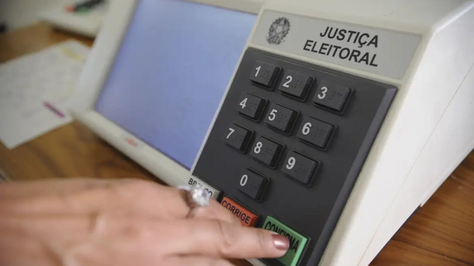 Mão pressiona botão "CONFIRMA" em urna eletrônica da Justiça Eleitoral, com teclado numérico