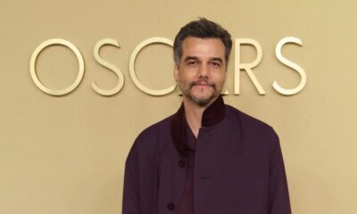 Wagner Moura, com barba e casaco roxo, em frente a um fundo dourado com o letreiro OSCARS