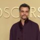 Wagner Moura, com barba e casaco roxo, em frente a um fundo dourado com o letreiro OSCARS