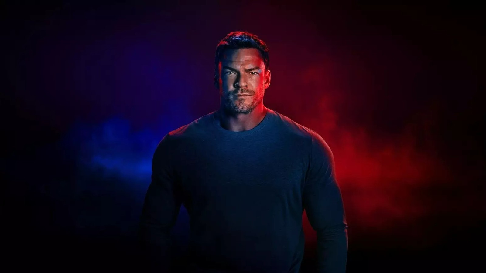 Homem de barba, camisa escura, olhando para frente, com fundo azul e vermelho esfumaçado