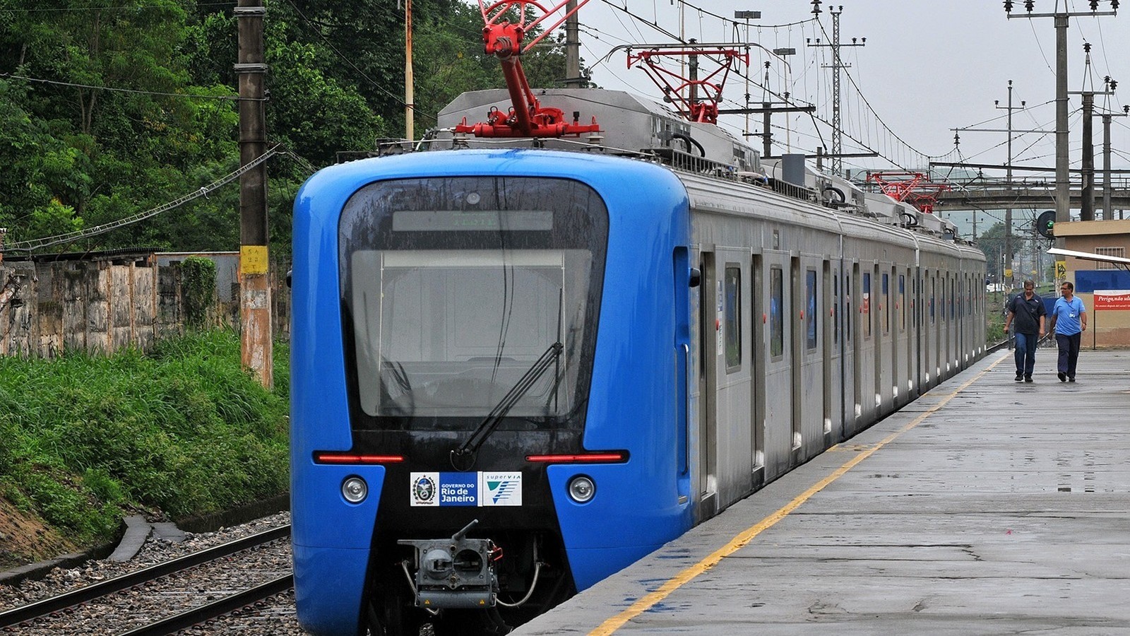 Trem azul e cinza da SuperVia em plataforma molhada, com dois homens caminhando, em dia nublado