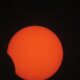 Sol laranja com manchas escuras, parcialmente coberto por um objeto circular escuro