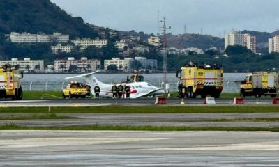 Avião branco fora da pista, cercado por bombeiros e veículos de emergência em aeroporto costeiro