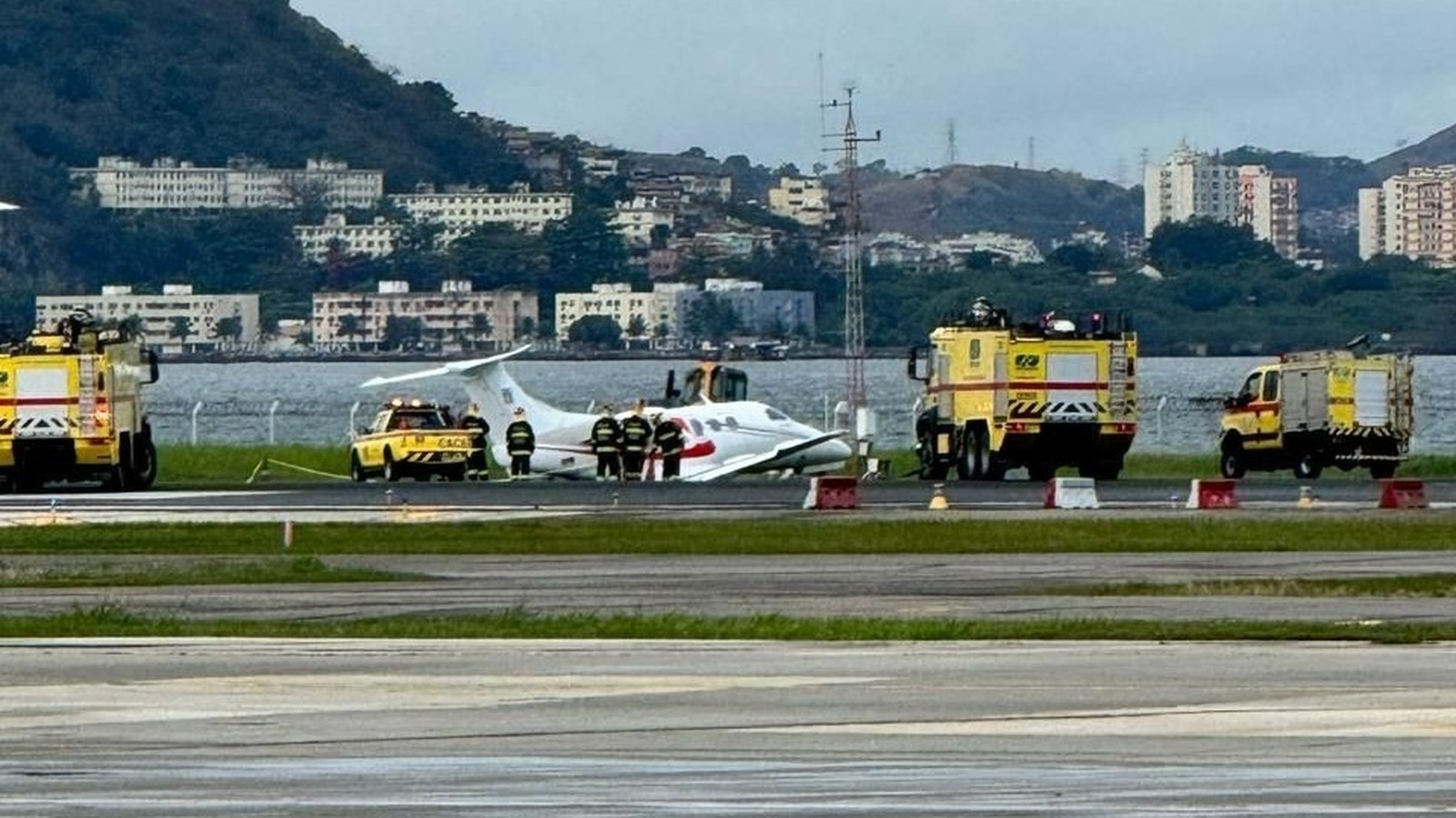 Avião branco fora da pista, cercado por bombeiros e veículos de emergência em aeroporto costeiro