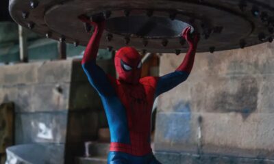 Homem-Aranha com traje vermelho e azul, erguendo grande objeto metálico acima da cabeça
