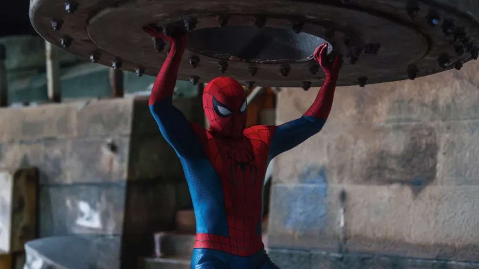 Homem-Aranha com traje vermelho e azul, erguendo grande objeto metálico acima da cabeça