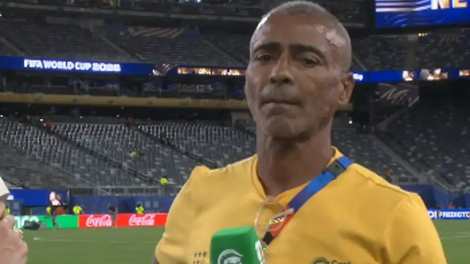 Homem de camisa amarela Romário com microfone verde em estádio, telão 'FIFA World Cup'