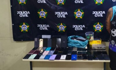 Dezenas de celulares e caixas sobre uma mesa, com um banner da Polícia Civil ao fundo