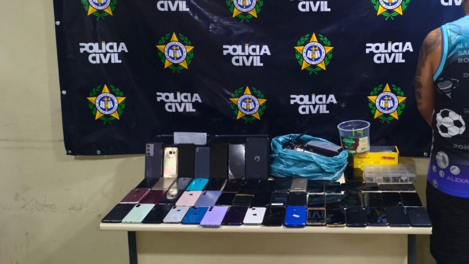 Dezenas de celulares e caixas sobre uma mesa, com um banner da Polícia Civil ao fundo