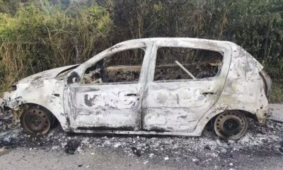 Carro hatchback totalmente carbonizado na lateral de uma estrada de asfalto, com mato ao fundo