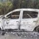 Carro hatchback totalmente carbonizado na lateral de uma estrada de asfalto, com mato ao fundo