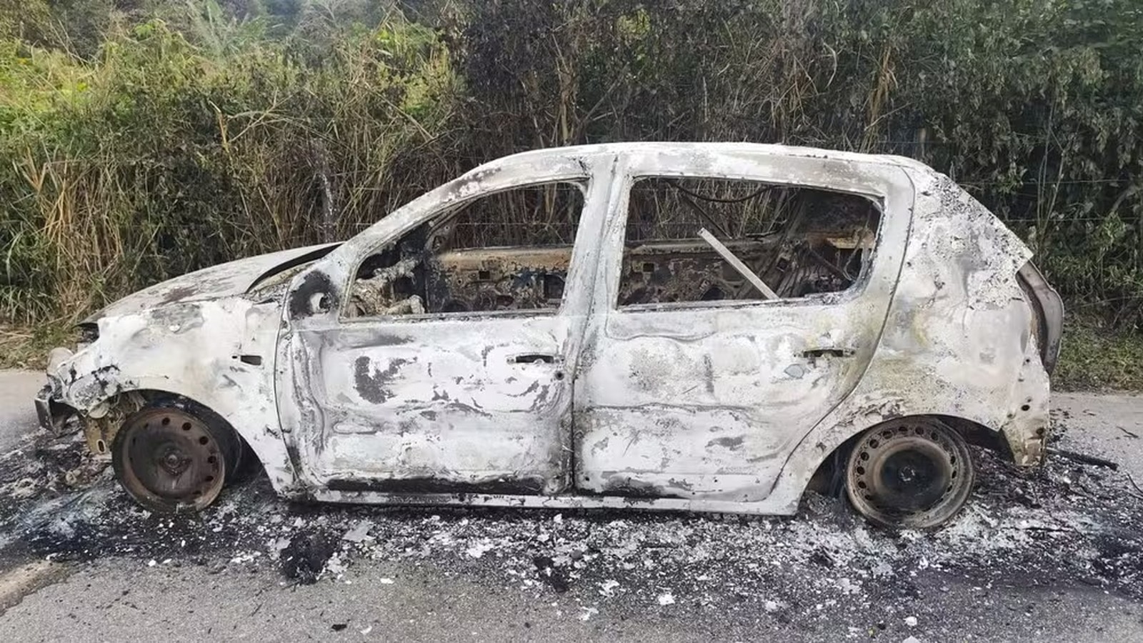 Carro hatchback totalmente carbonizado na lateral de uma estrada de asfalto, com mato ao fundo