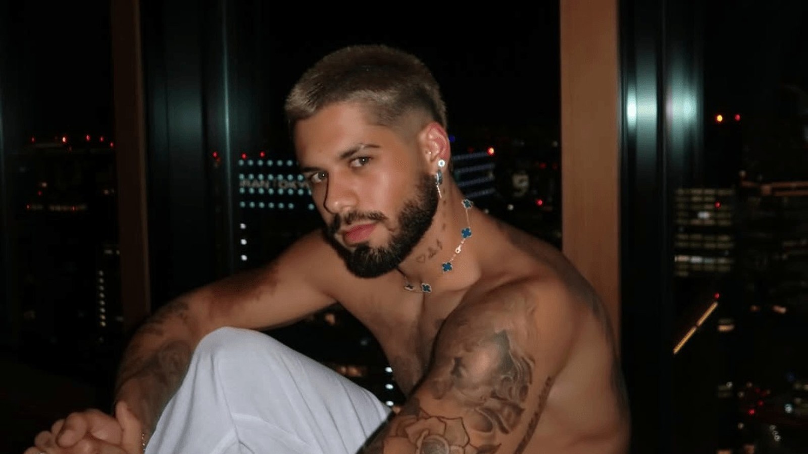 Zé Felipe sem camisa, com barba e colar, vista noturna ao fundo