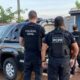 Três policiais civis de costas com coletes 'PCPR' e 'POLÍCIA CIVIL', viatura e carro