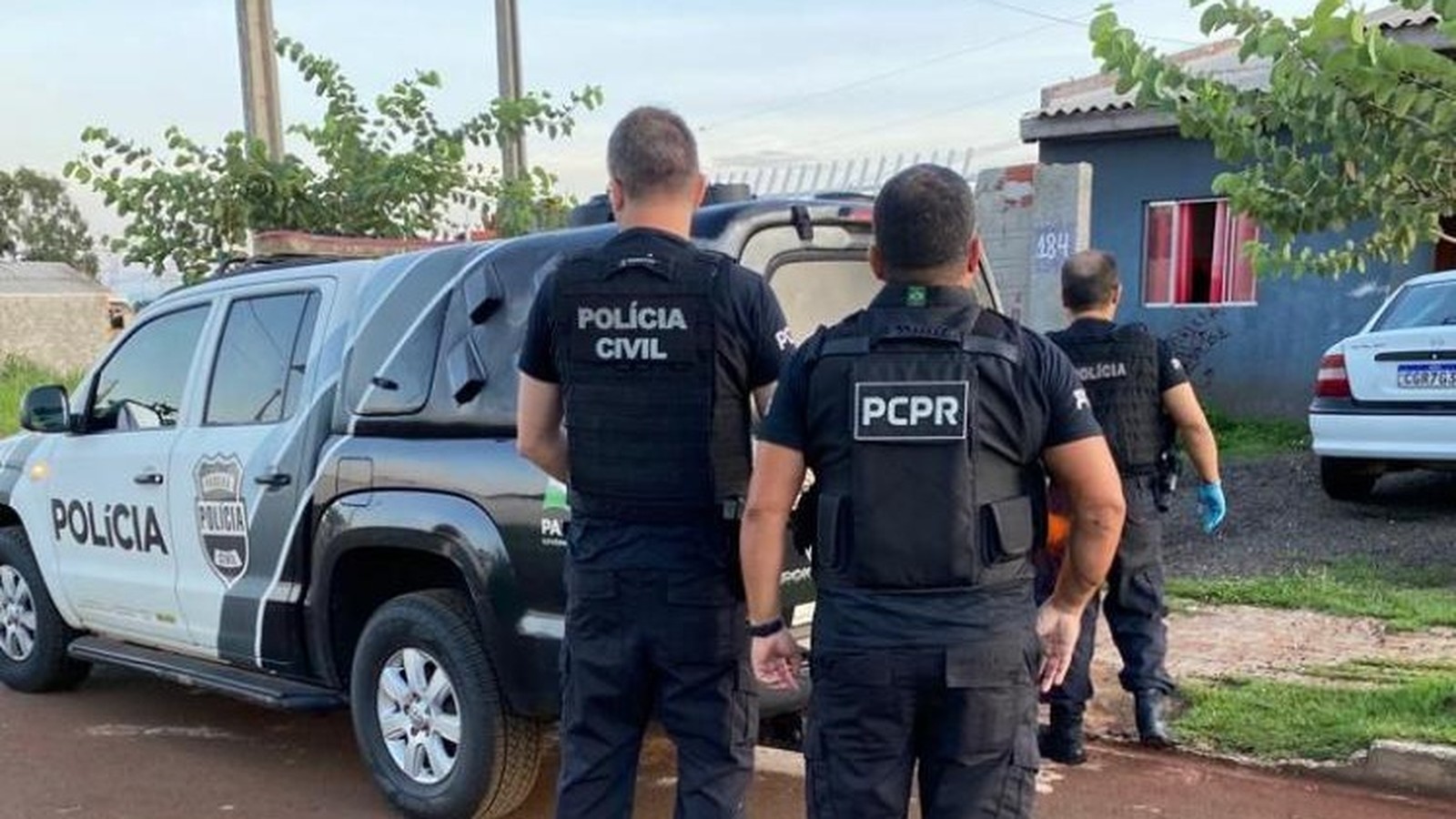 Três policiais civis de costas com coletes 'PCPR' e 'POLÍCIA CIVIL', viatura e carro
