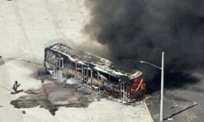 Visão aérea de ônibus carbonizado em chamas com fumaça escura e dois bombeiros o combatendo