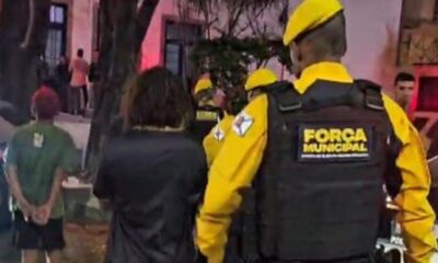 Agentes da Força Municipal em uniformes amarelos escoltam indivíduos à noite, um algemado