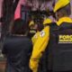 Agentes da Força Municipal em uniformes amarelos escoltam indivíduos à noite, um algemado