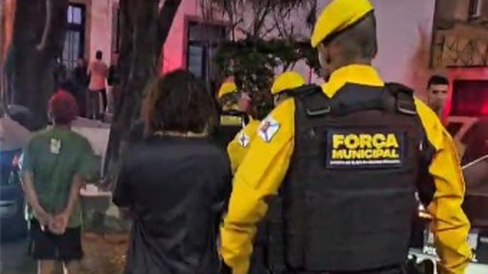 Agentes da Força Municipal em uniformes amarelos escoltam indivíduos à noite, um algemado