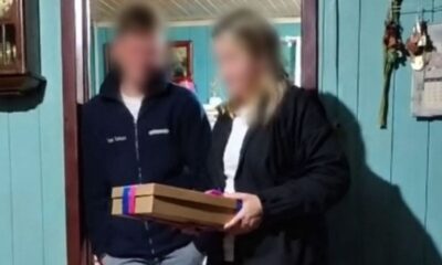 Duas pessoas borradas, homem e mulher, segurando uma caixa marrom com fita rosa e azul em uma porta