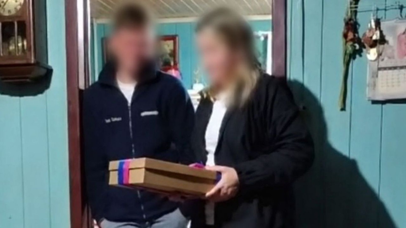 Duas pessoas borradas, homem e mulher, segurando uma caixa marrom com fita rosa e azul em uma porta
